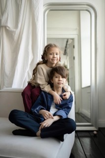 A brother and sister sit together on a white sofa in a bright, minimalistic room The siblings share
