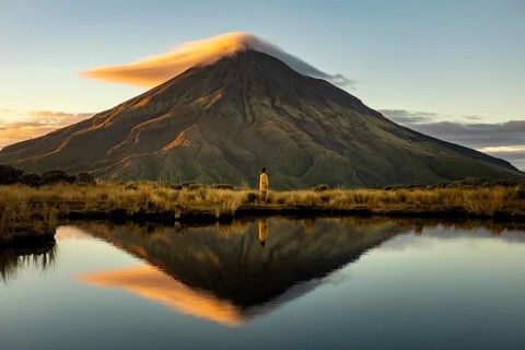 Side view of unrecognizable person standing by a reflective lake, gazing at the majestic Mount