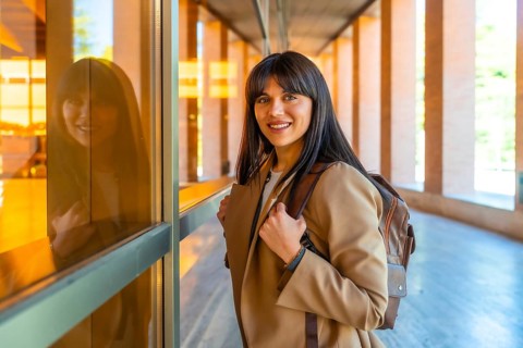 Young woman carrying a backpack and walking along a modern university campus building, smiling