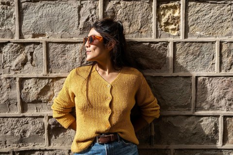 Latin woman in trendy sunglasses leans against a textured stone wall, basking in warm sunlight. She
