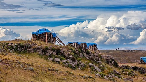 Wooden structures covered by tarps on a hill side under cloudy sky, The Chullpas of Sillustani near