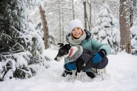 A smiling child in winter attire hugs a dog, both enjoying a snowy forest day. The scene captures
