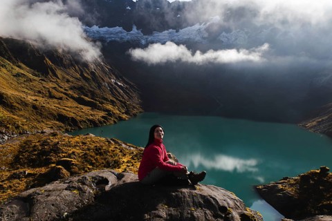 Ecuadorian woman in a red sweater sits on a rock facing a tranquil turquoise lake and lush
