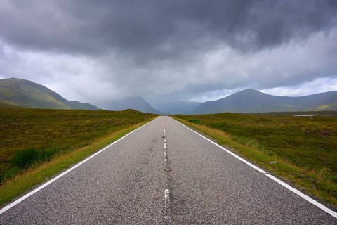 A long, straight road leads through a vast landscape of low-hanging clouds and mountains, Rannoch