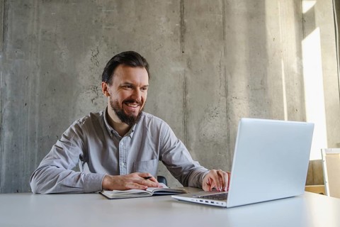 A professional man sits smiling at a desk, engaging with his laptop while taking notes. Natural