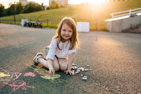 A child sits on pavement, happily drawing with colorful chalk in a sunny park. The scene captures a