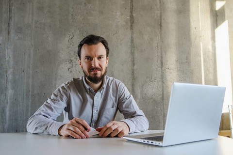 A confident man sits at a desk with a laptop, exuding professionalism. The concrete wall background