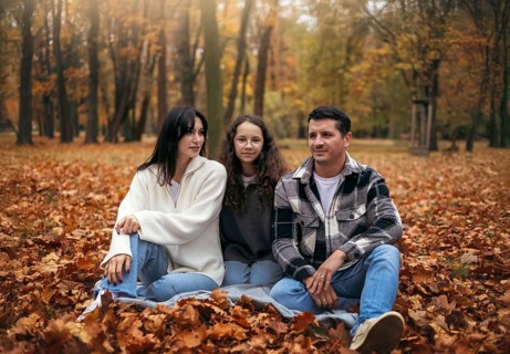 A family sits together among fallen leaves on a crisp autumn day in a tree-filled park. They are