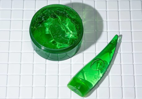 Open container of vibrant green aloe gel next to a body care cream tube on a white tiled bathroom