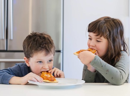 Two young siblings joyfully eat pizza in a modern kitchen, showcasing the simplicity of childhood