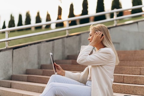 A businesswoman in a white outfit sits on outdoor stairs, holding a tablet. She appears engaged,