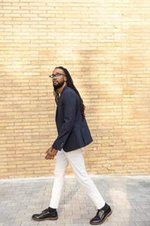 A cuban businessman with long hair in a navy blazer and white pants walks confidently past a brick