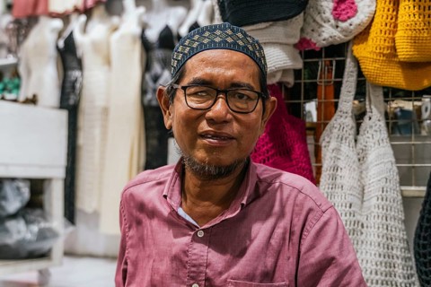 A balinese man in a pink shirt smiles while standing in front of colorful crocheted bags at a