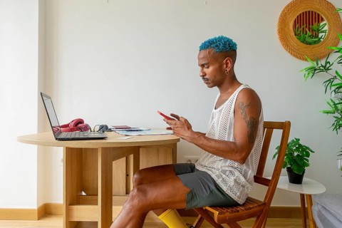 A fashionable LGBTQIA+ man with blue hair sits at a wooden table, focused on a phone, planning