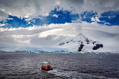 A small boat sails across the sea against a dramatic backdrop of snow-covered mountains and clouds,