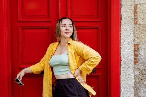 A confident woman poses stylishly in front of a vibrant red door, wearing a bright yellow shirt and