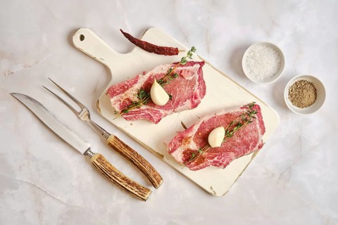 Two raw pork rib chops are placed on a wooden cutting board. Garlic cloves and fresh herbs are