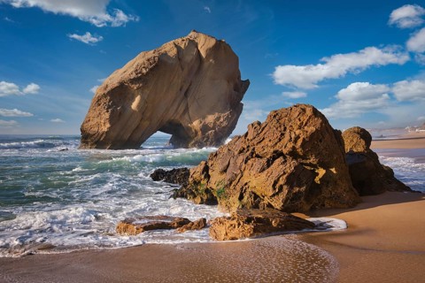 Sunset casts a warm glow on Penedo do Guincho rock arch, framed by the surf and sandy beach at
