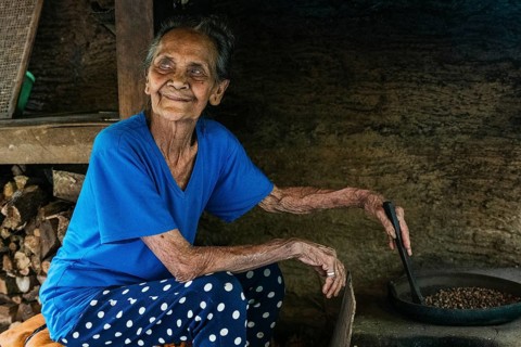 An elderly woman in a blue shirt and polka dot pants is roasting balinese coffee beans in a rustic