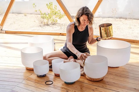 A woman is seated on a wooden floor, playing multiple crystal singing bowls. She gently taps them