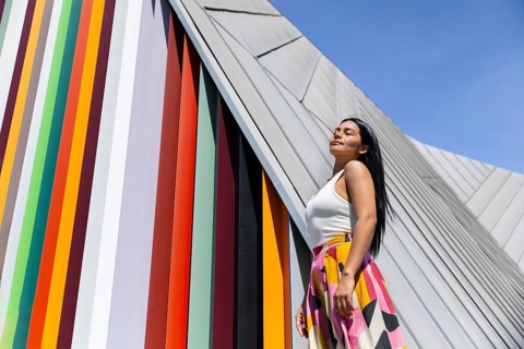 A woman in a vibrant outfit stands confidently against a backdrop of modern architecture. Her