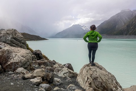 A woman in a green jacket stands on a rocky outcrop, overlooking the serene landscape of Mount Cook