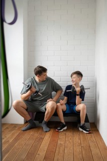 A father and son bond while exercising with dumbbells at home, sitting on a bench in a bright room.