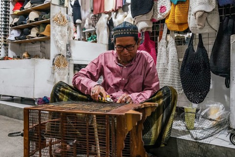 A balinese artisan skillfully crafting a product, surrounded by colorful handmade crochet bags in a