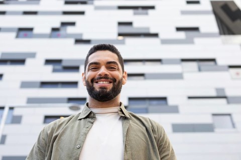 A man with a beard smiles confidently, standing before a modern, geometric patterned building. The