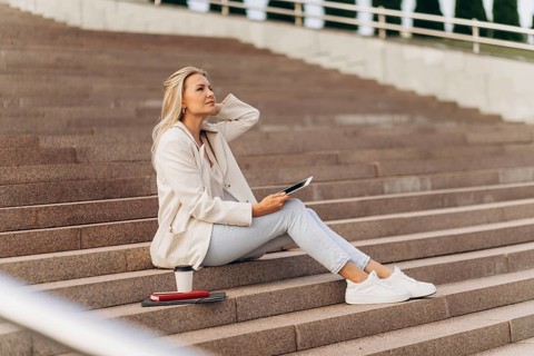 A businesswoman in casual attire sits thoughtfully on steps holding a tablet, with a coffee cup and