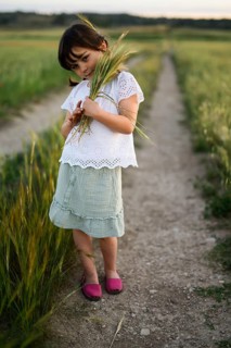A young girl, dressed in a white blouse and green skirt, gently holds a bunch of wheat while