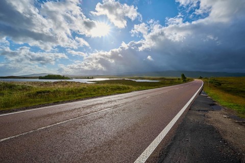 Long road with beautiful sunlight, flanked by greenery and expanses of water, cloudy on the