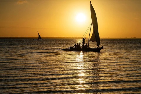 Silhouette of a traditional dhow sailing boat with people on board against a golden sunset over the