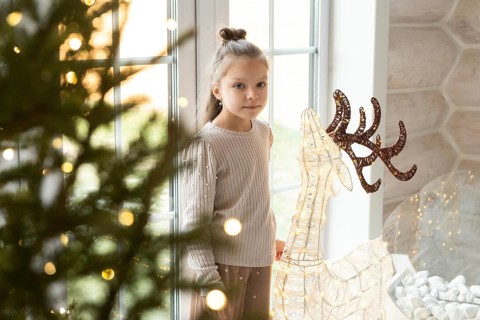 A young girl stands by a window adorned with Christmas lights, looking at camera, next to a