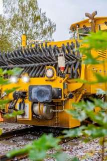 Yellow machine on rails, surrounded by foliage, showing technical components, tamping machine,