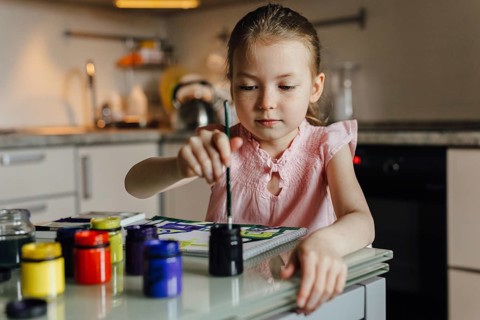 A young girl in a pink blouse paints attentively at a table in a cozy kitchen. Surrounded by