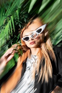 A fashionable woman wearing white cat-eye sunglasses stands amidst lush green foliage. The photo