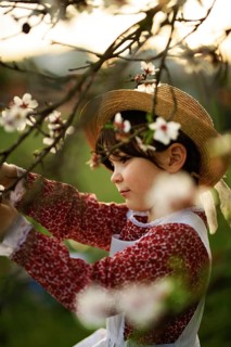 A young girl in a red dress and straw hat admires the blossoms of an almond tree. The scene