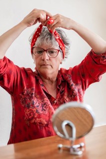 A senior woman with gray hair and glasses ties a red polka dot headscarf in front of a mirror.