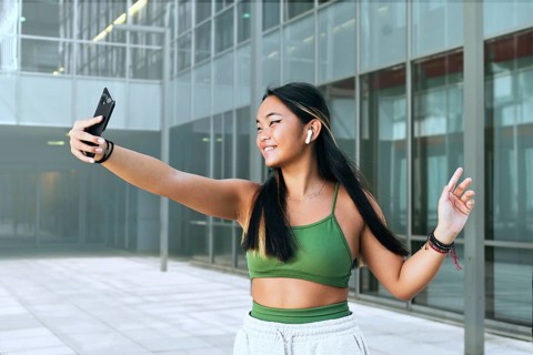 A Chinese teenager wearing wireless earbuds and a green top takes a selfie outdoors. She is in a