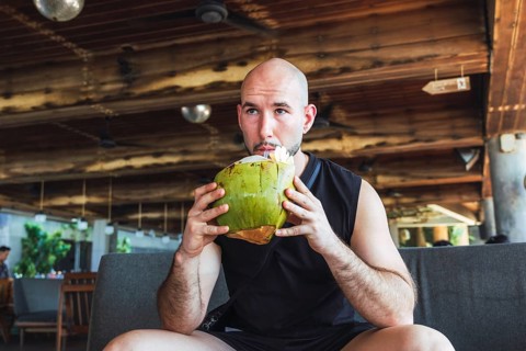 A bald man sips from a green coconut while relaxing indoors on his vacation in Bali. This casual,