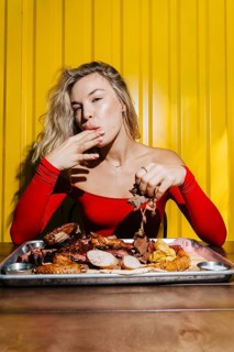 A woman in a vibrant red outfit relishes assorted barbecued meats on a tray. The vibrant yellow