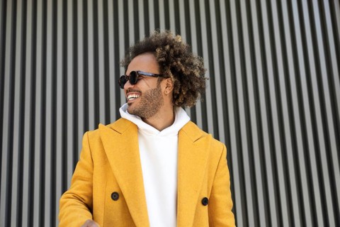 An afro man wearing a vibrant yellow coat and sunglasses, standing confidently against a modern,