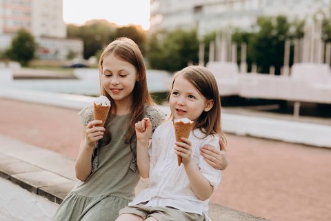 Two young girls sit on a ledge, enjoying ice cream cones on a sunny day. Their cheerful expressions