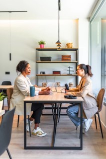 Two businesswomen are recording a podcast in a modern office, using a microphone and laptop