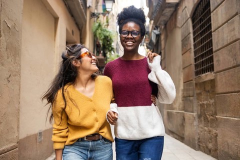 Two friends walk arm in arm down a charming street, laughing joyfully. One wears sunglasses, adding