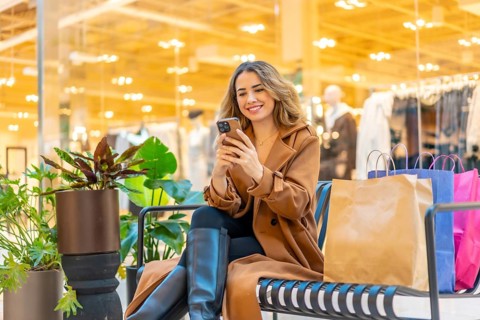 Woman in a trench coat sitting on a bench, looking at her phone and smiling, with shopping bags