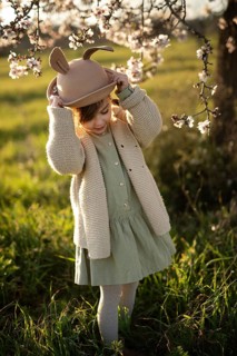 A young girl in a whimsical rabbit hat adjusts her hat while standing amidst beautiful almond