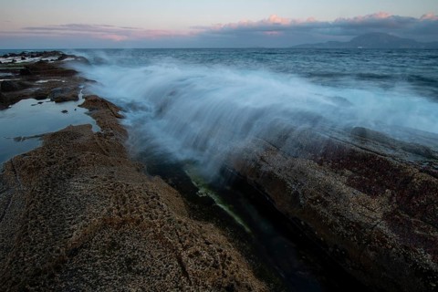 Waves crash against rocky shores under a vivid sunset sky in Tarifa, Cadiz. The vibrant colors and