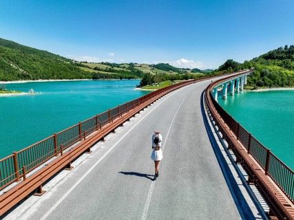 A young woman walks along a picturesque bridge overlooking a vibrant turquoise lake, surrounded by
